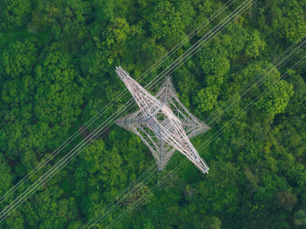 Pohľad zhora na veľký elektrický stĺp obklopený telenými stromami.